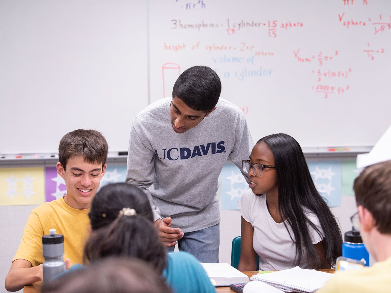 A student wearing a light gray long-sleeve UC Davis shirt leans down to assist two junior high school students. In the background is a large whiteboard with equations written on it.