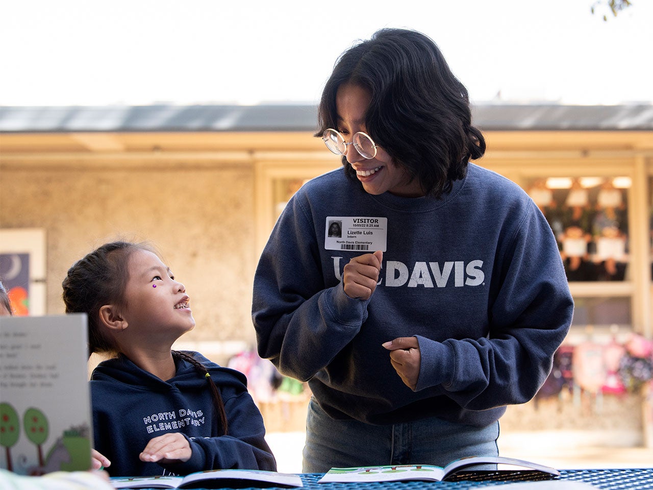 A student in a dark blue UC Davis sweater and round-rimmed glasses helps an elementary student read a picture book. The elementary student looks up at the UC Davis student with a big smile.