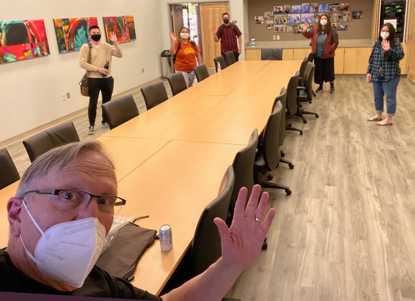 Professor (masked) gives a wave in selfie as masked students enter conference room.