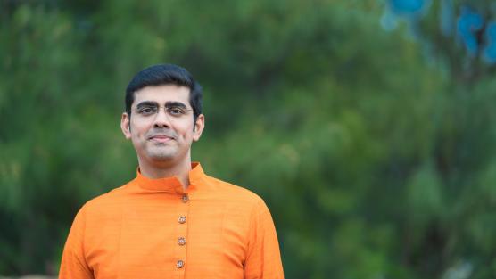 Musician named Charan standing left of center in a saffron-colored band collar shirt. The background is a green blurred forest.