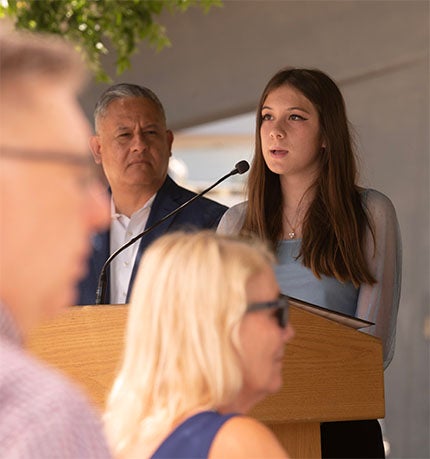 Young woman singing at a podium with a man standing behind, blurred audience in foreground