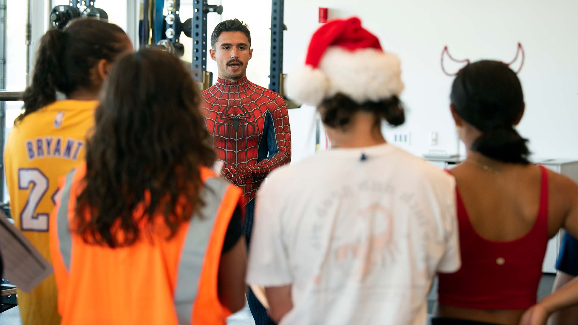 Coach in Spider-Man costume talks to student-athletes in Halloween costumes in weight room