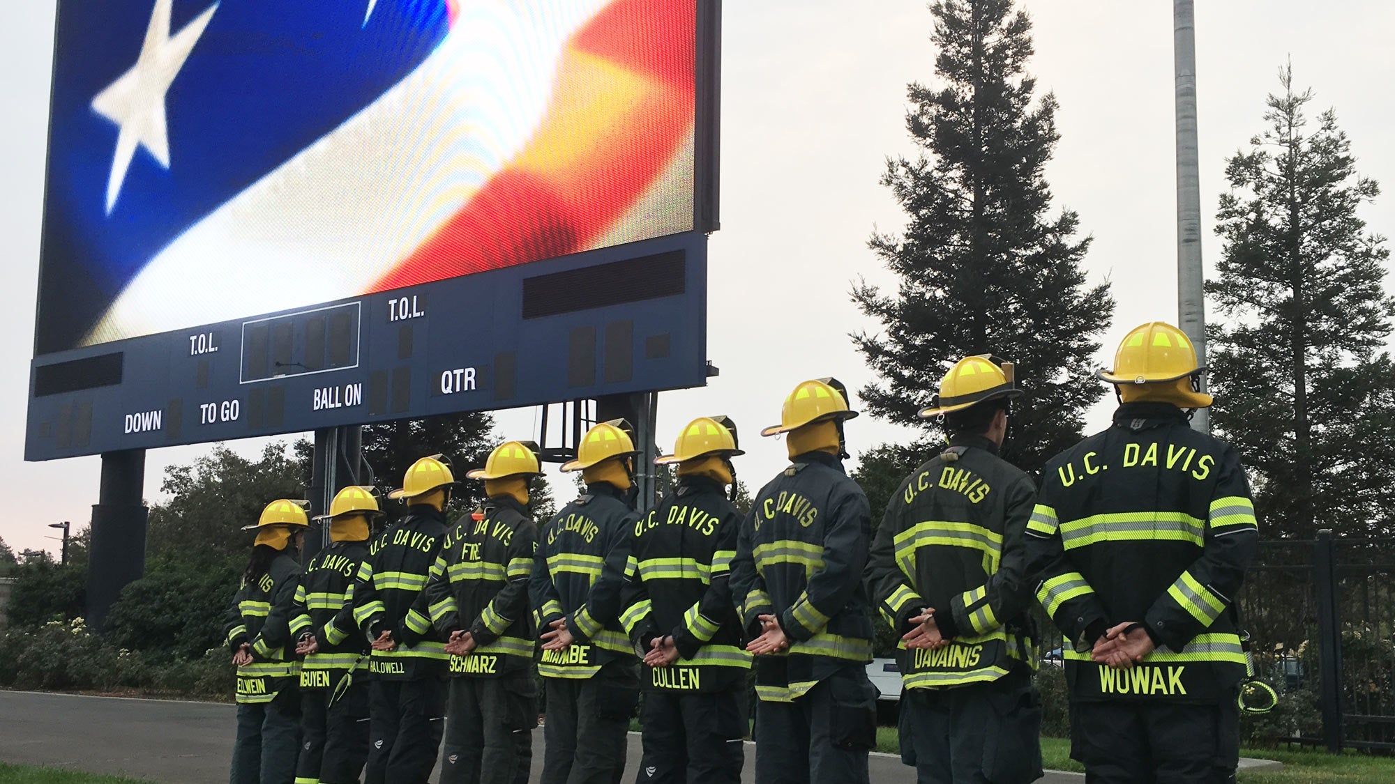 Firefighters in turnouts stand at attention for flag raising, under stadium scoreboard showing the Stars and Stripes