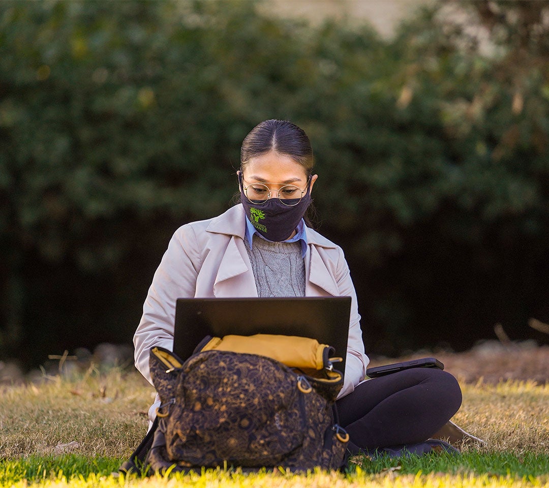 Student working on a laptop outdoors at UC Davis Arboretum, maintaining health protocols with a face mask.