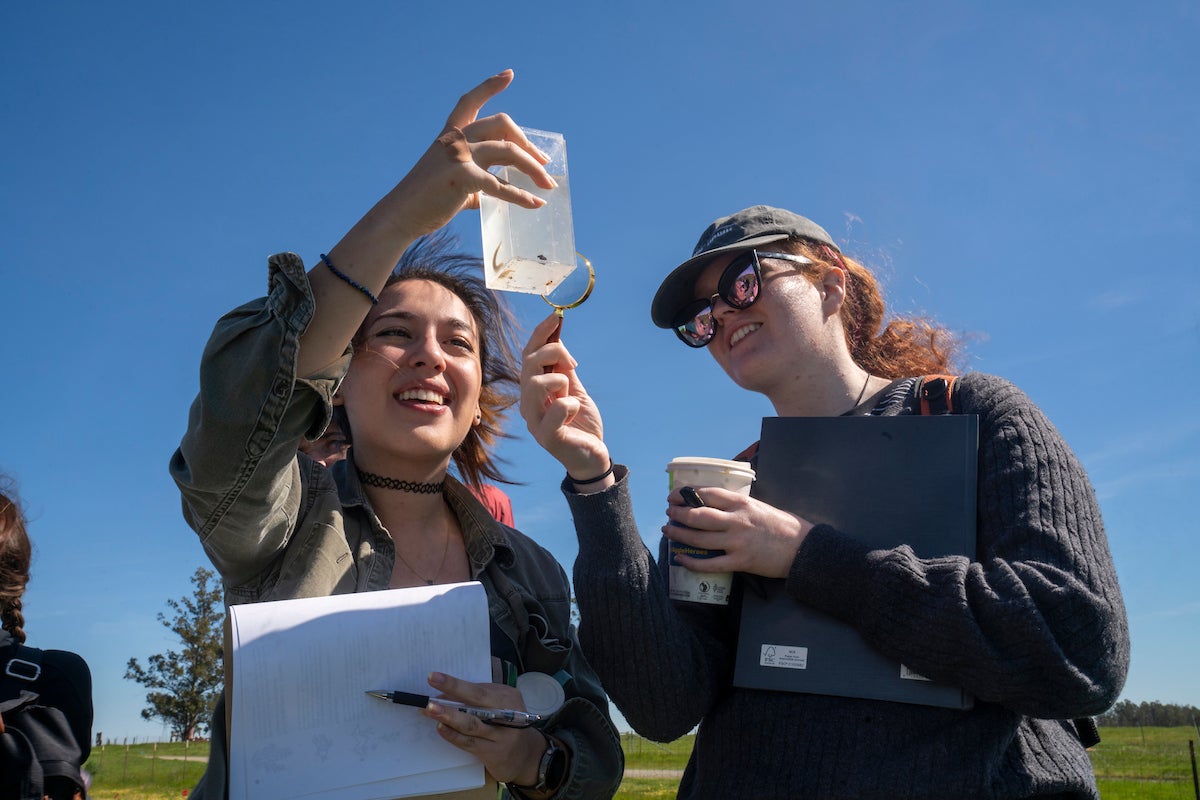 Two students smile into the sun and examine a sample. 