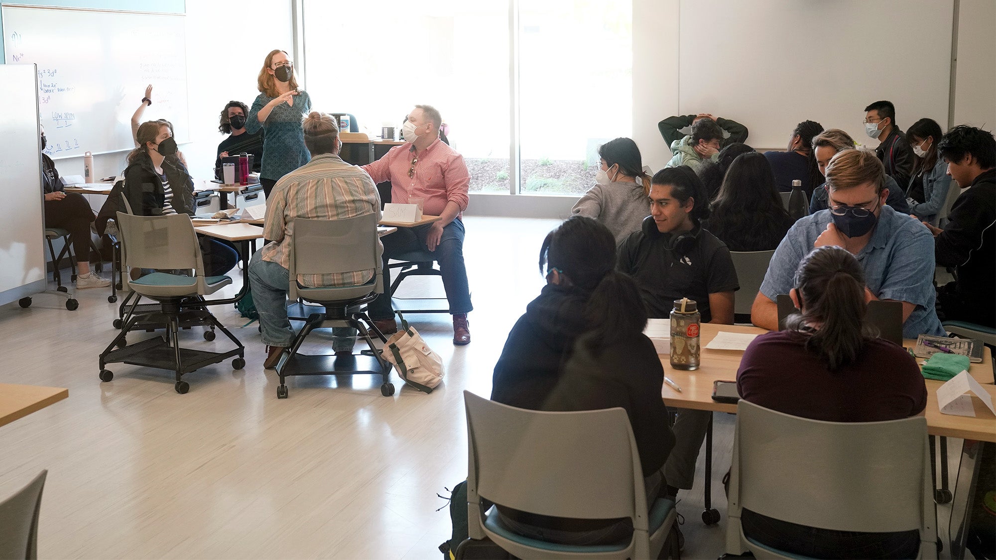 Instructor speaks while students in desks — some arranged in circles — listen.