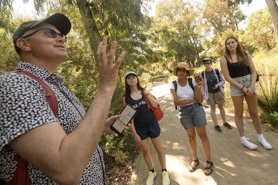 Smiling guide gesturing to five young adults on a sunlit park path