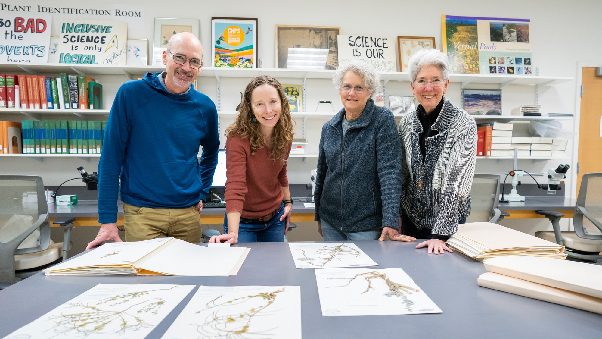 Four people smiling, leaning on a table with botanical prints in the forground.