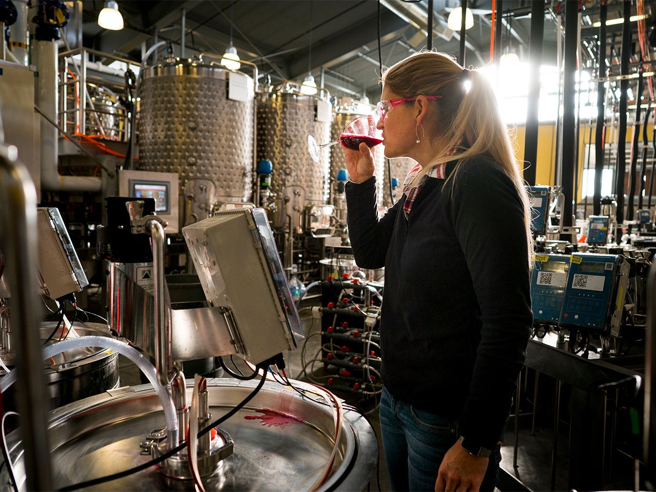 Anita, surrounded by metallic fermentation tanks and wearing safety goggles, tastes red wine from a wine glass.