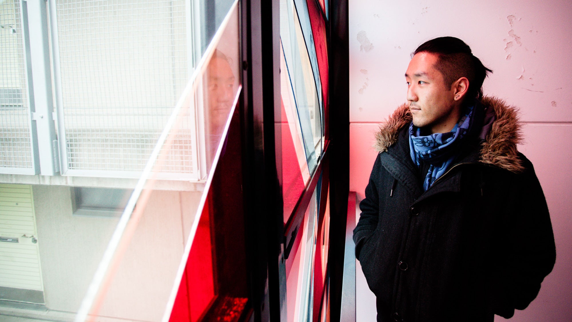 Man in black coat and blue scarf gazes through red-tinted window, photograph