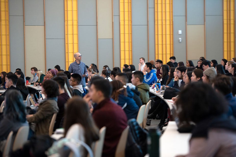  Lecturer walks amid rows of tables of students, while lecturing.
