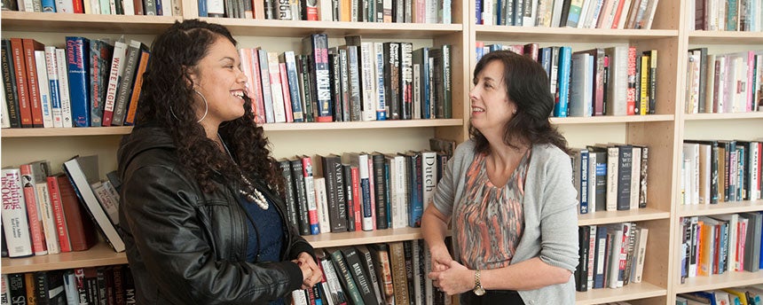 Two women standing against a book shelf talking