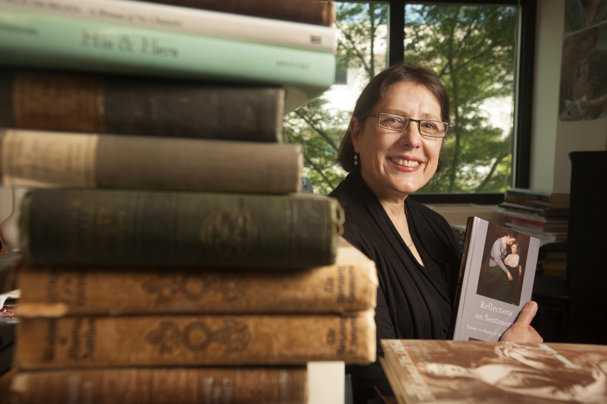 A female professor is pictured with a stack of books in the foreground