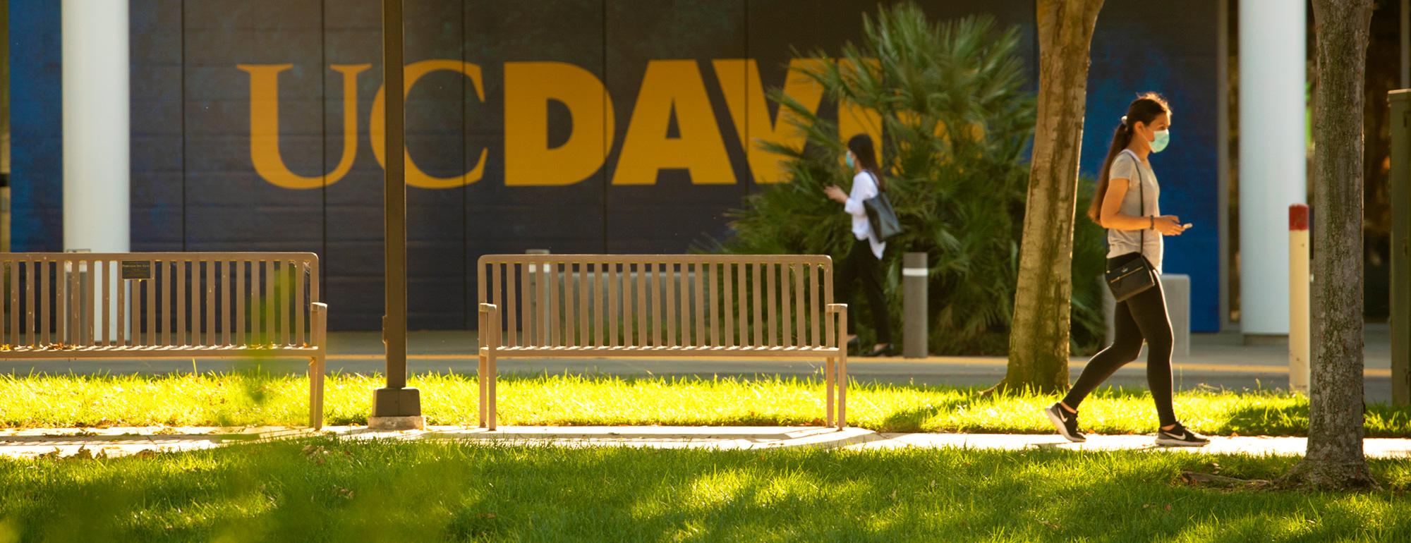 A student walking on campus. A large UC Davis logo on a building wall in the background.