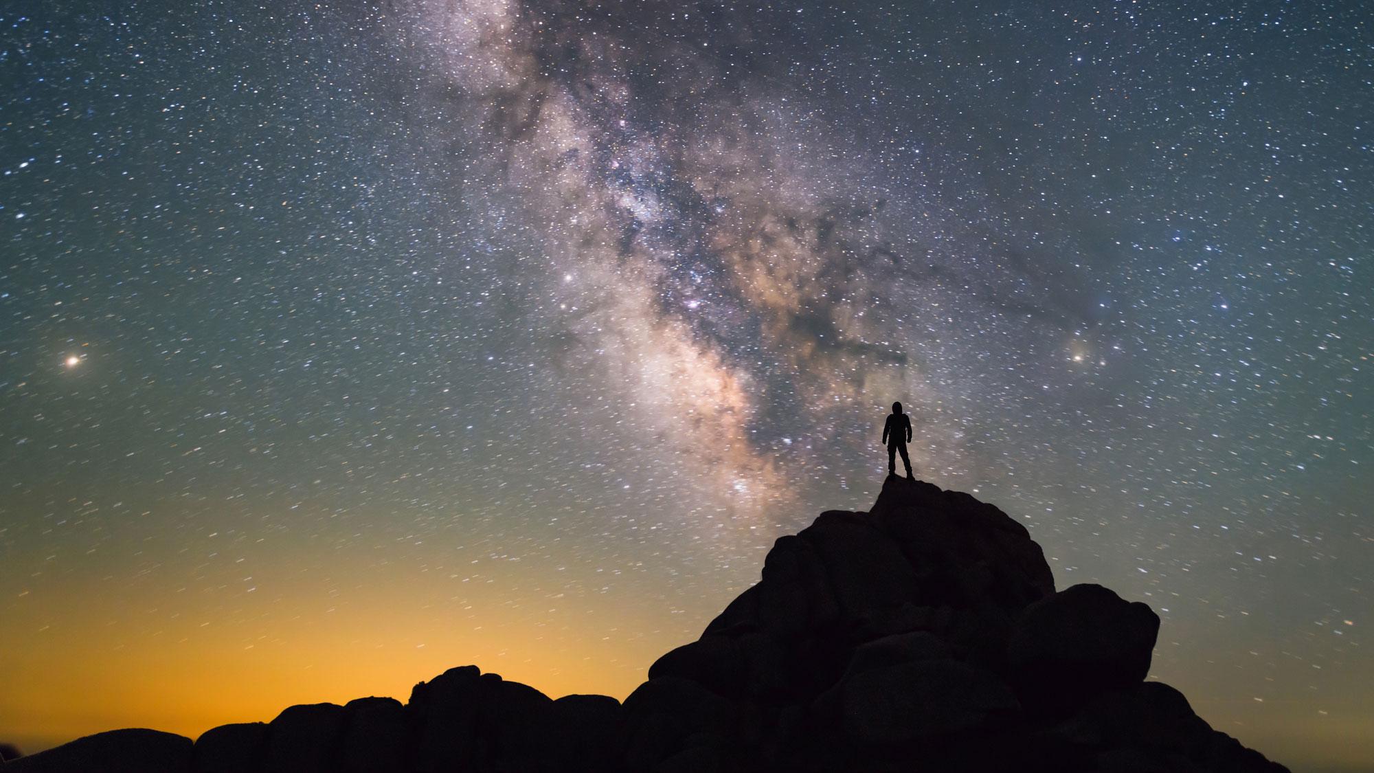 A person stands atop a small mountain in the foreground facing a star filled sky