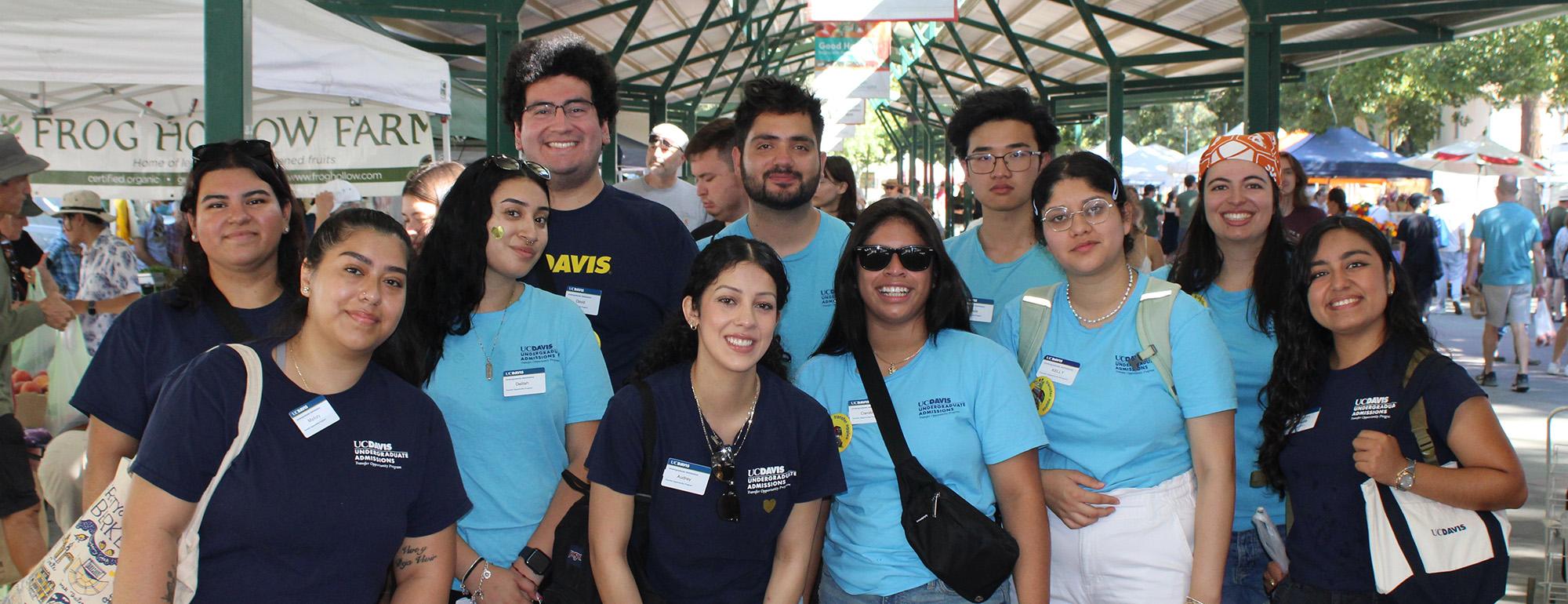 Diverse group of smiling volunteers in blue shirts under a canopy at an outdoor fair