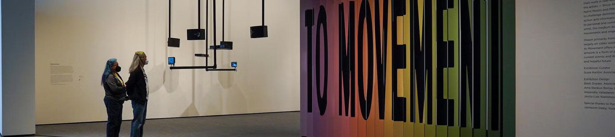 Gallery with two visitors viewing hanging black panels and colorful 'TO MOVEMENT' text