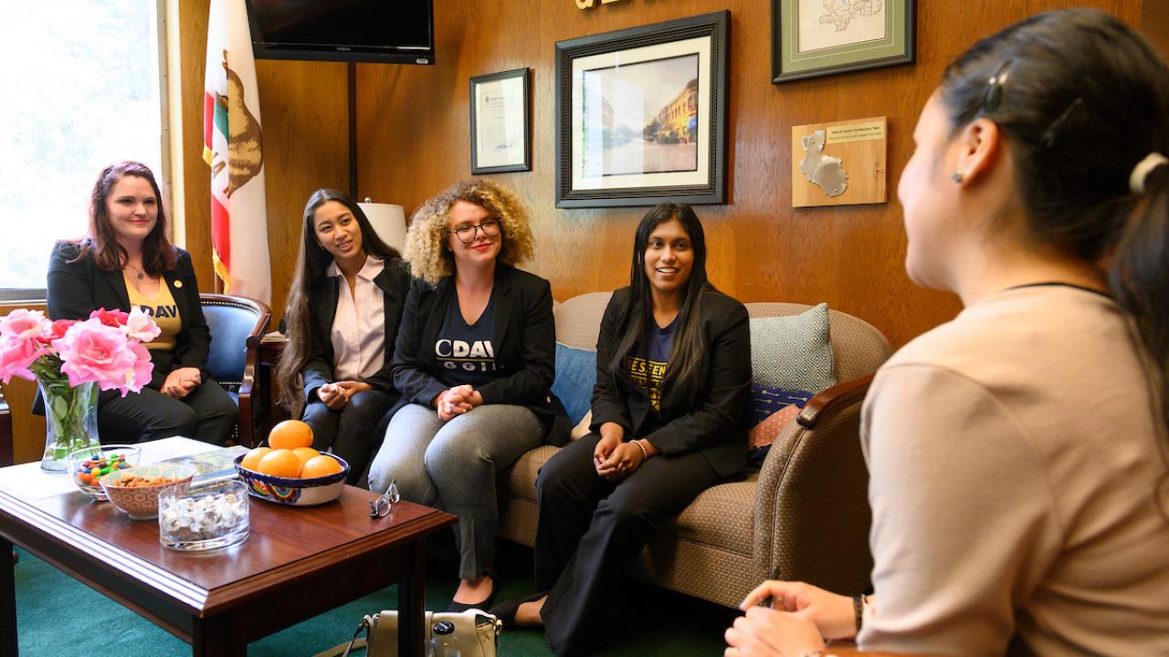 Four students sit on a couch in an office with flowers and a bowl of oranges while a politician listens.