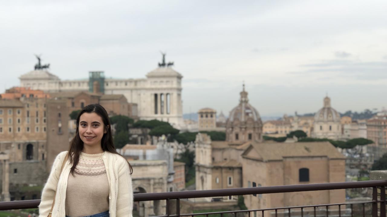 Katharine wears a cardigan and stands in front of a railing and a city scape with domes and buildings of various heights. 