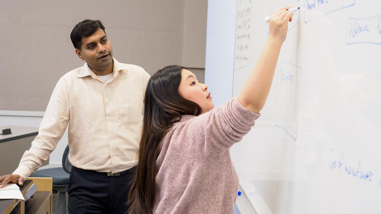 Business school student YiYi Han (foreground) at the white board writing while an instructor watches from behind.