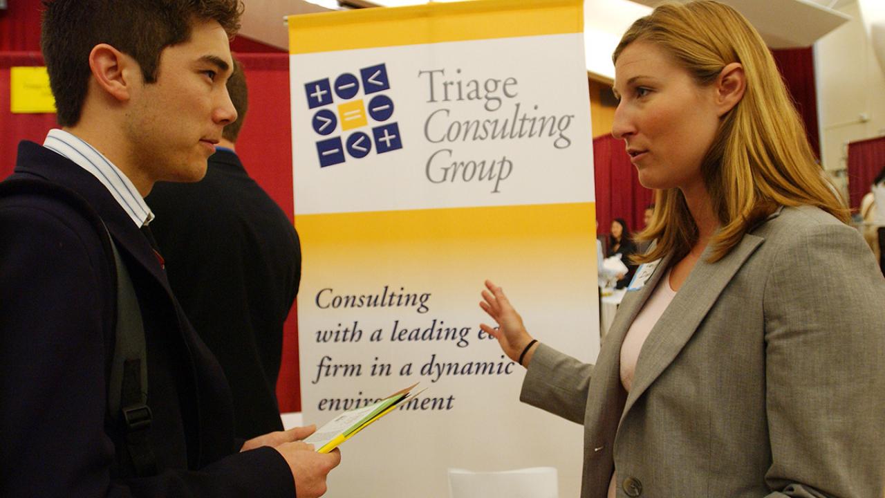 A male student listening attentively to a female representative of Triage Consulting Group at a career fair