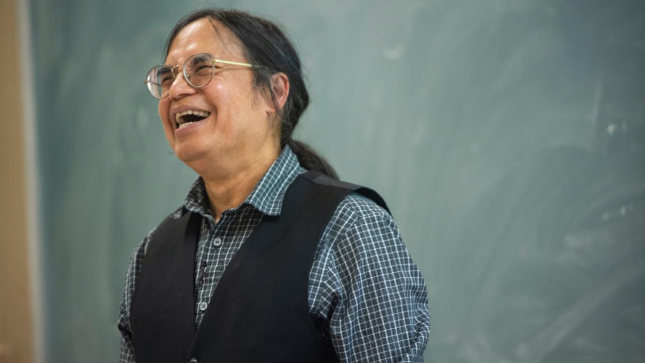 Male professor laughs in front of blackboard