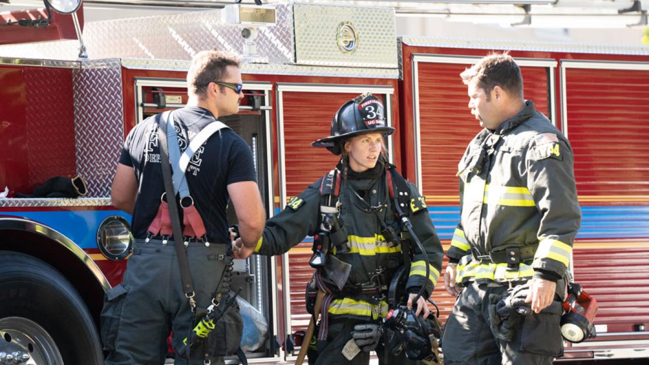 Three firefighters, in gear, in front of firetruck