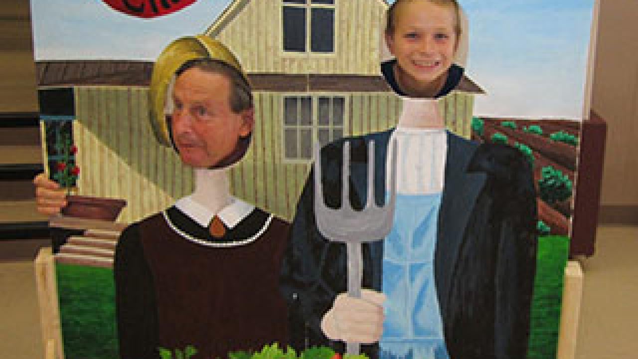 Man and boy stick their heads through a cut-out board with the American Gothic as the theme