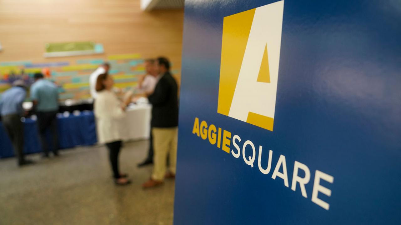 "Aggie Square" logo sign in foreground of meeting.