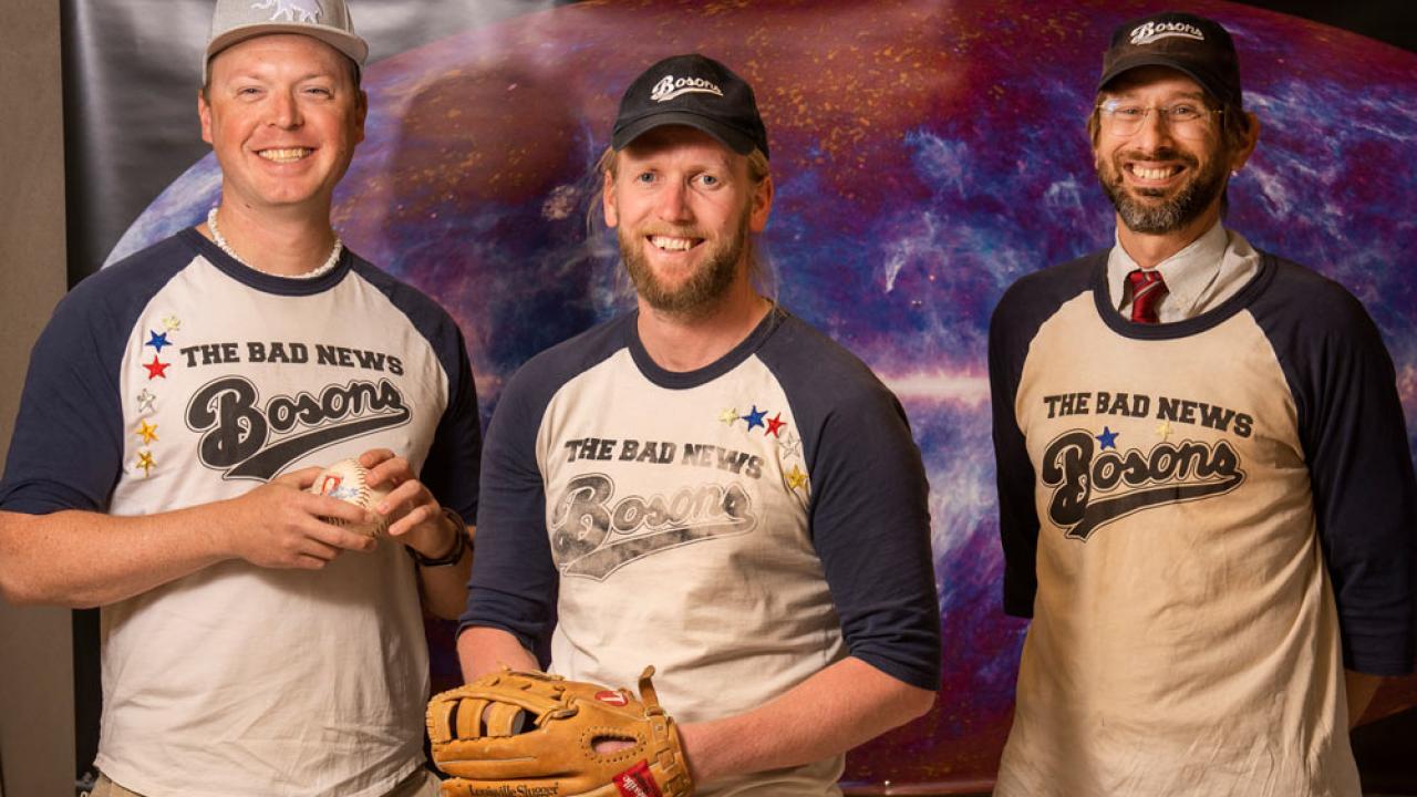 Three men, softball players, in "The Bad News Bosons" jerseys. One man holds a softball, another has a glove on.