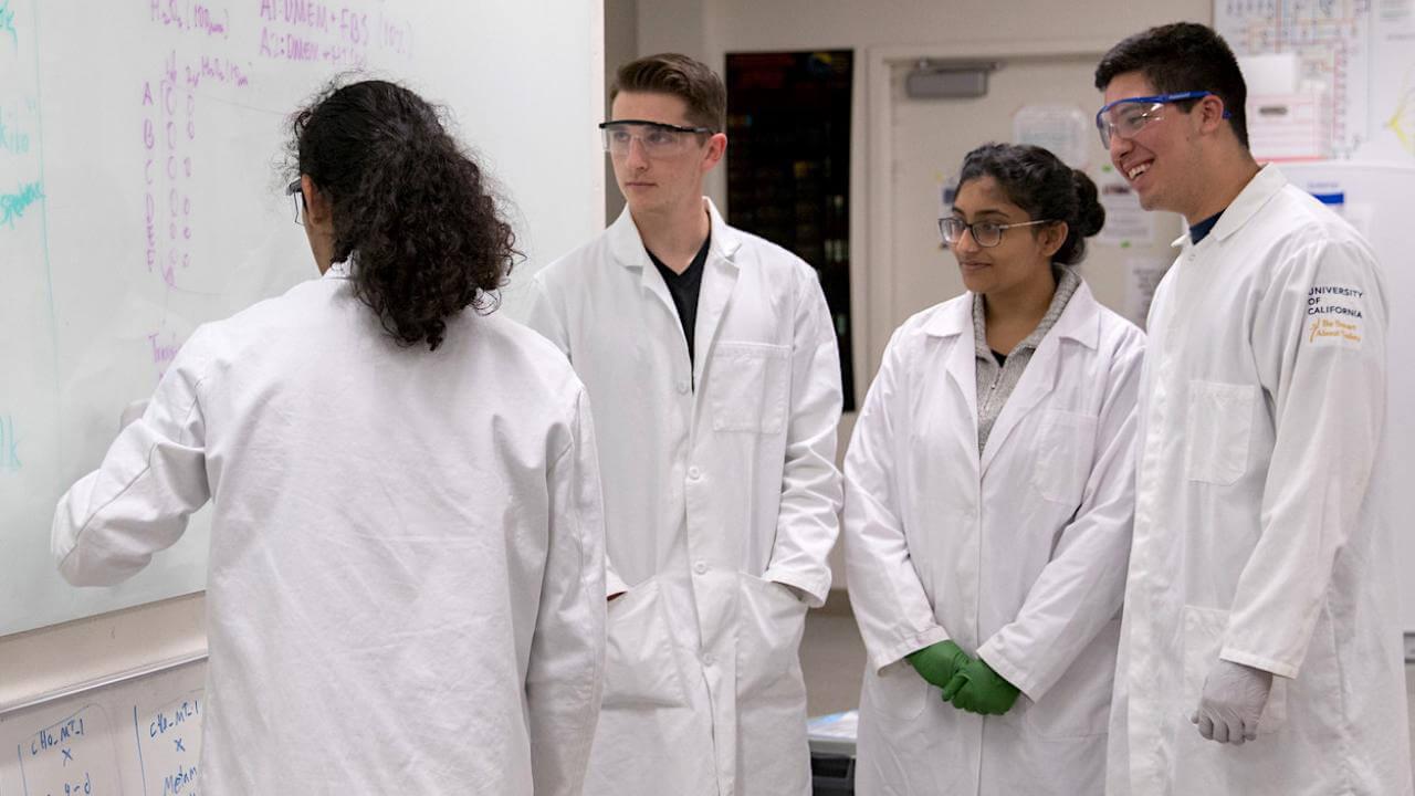 Students in lab coats stand in front of a white board.