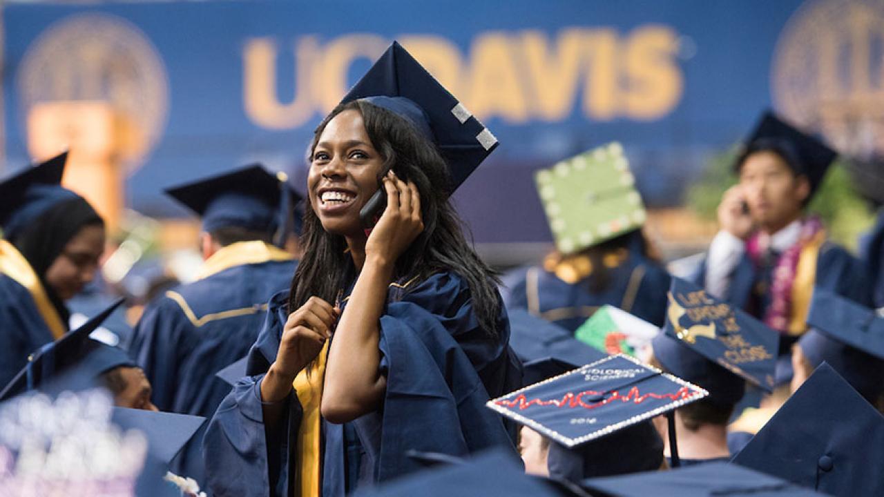 In a sea of other graduates, a female in graduation cap and gown uses her cell phone