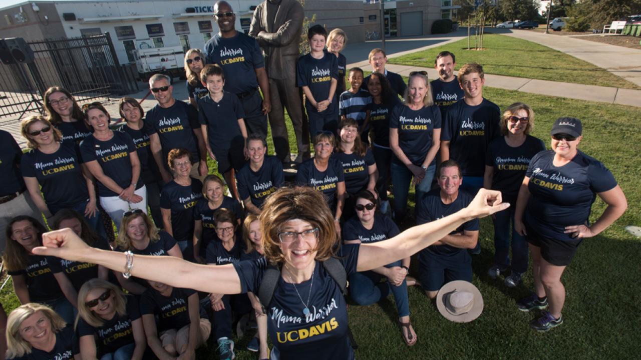 Janice Corbett, arms outstretched, with Warriors behind her, around Jim Sochor statue outside Aggie Stadium