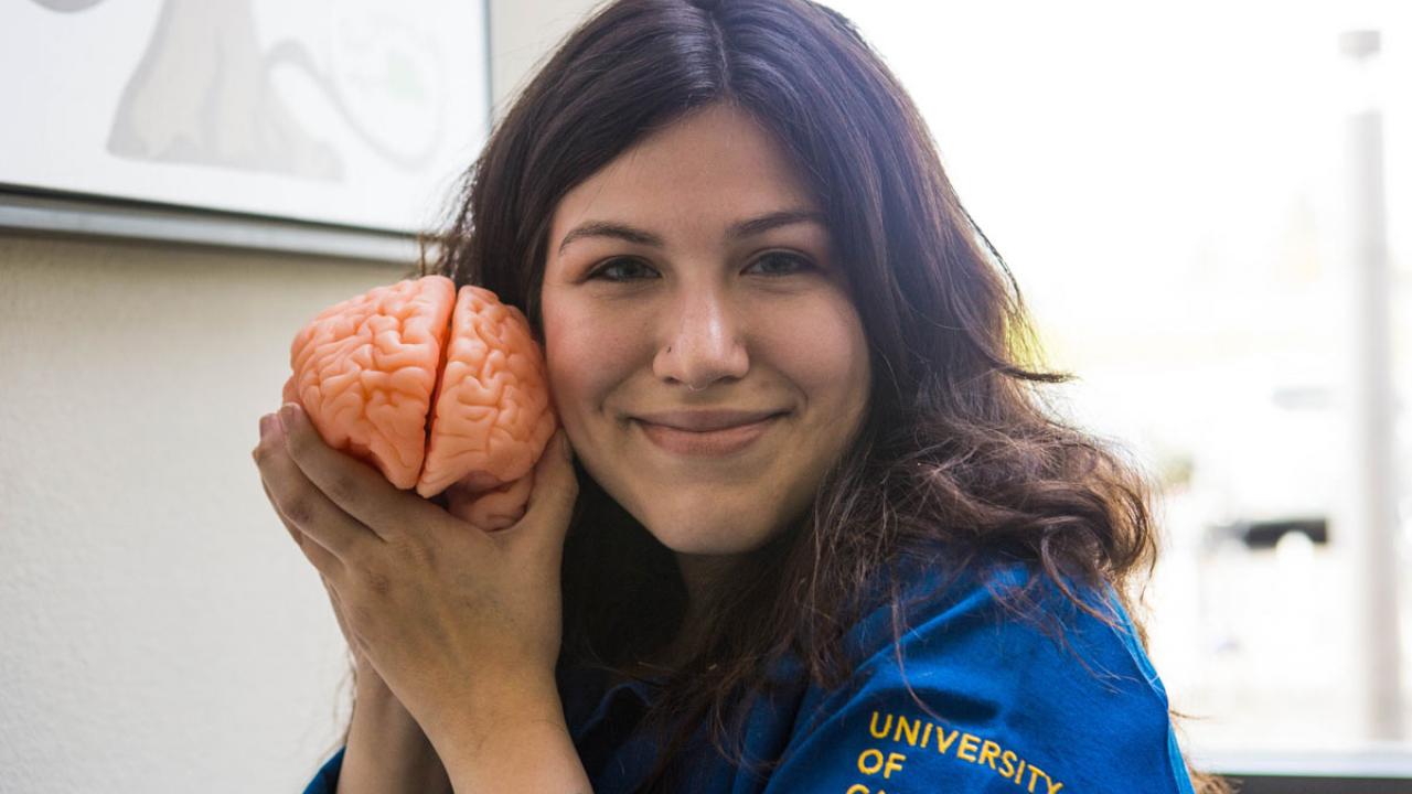 Female student  in UC Davis blue lab coat holds model of brain close to her smiling face.