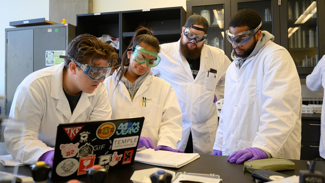 Four students wearing lab coats, goggles and gloves stand near a laptop.