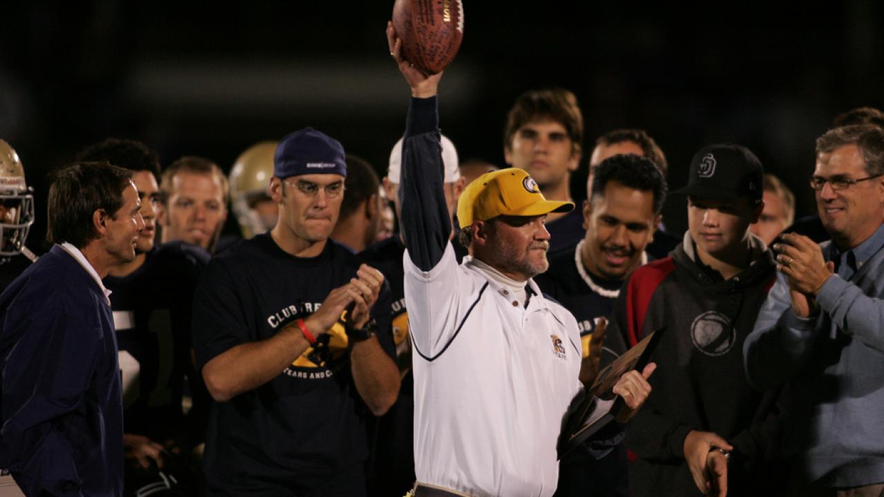 Coach Fred Aro holds football in air, as if in triumph, on sidelines.