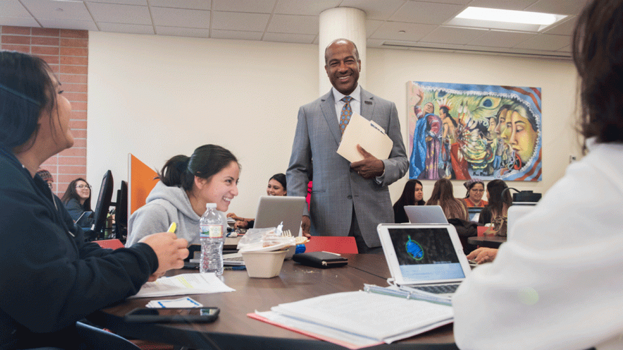 Chancellor Gary S. May, standing, talking, at a table full of students.