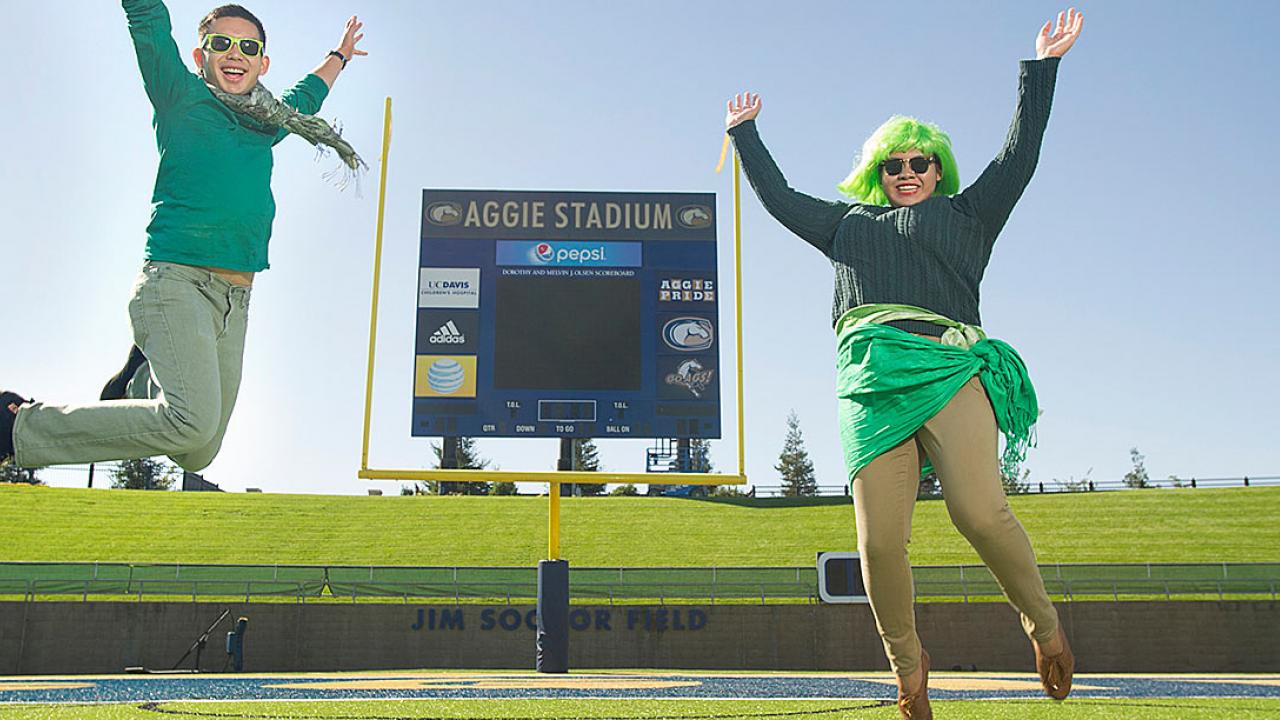 Photo: A woman and a man dressed in several pieces of green clothing pose in front of the Aggie Stadium scoreboard