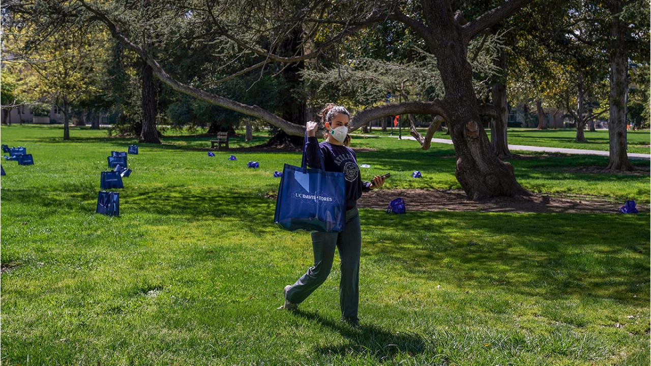 Wioman leaves Quad with a bag of groceries, from an array of bags set 6 feet apart on the Quad.