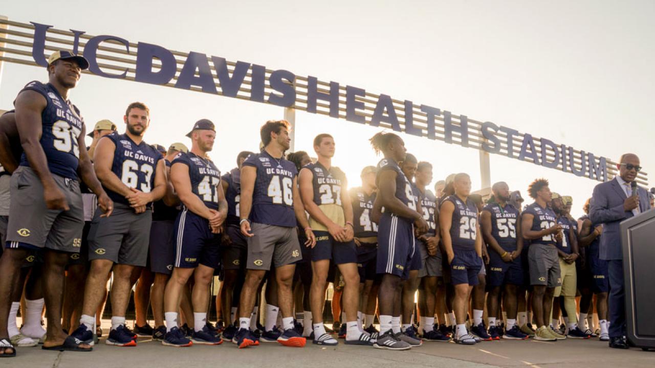 Student-athletes stand under new "UC Davis Health System" sign while Chancellor May talks at podium.