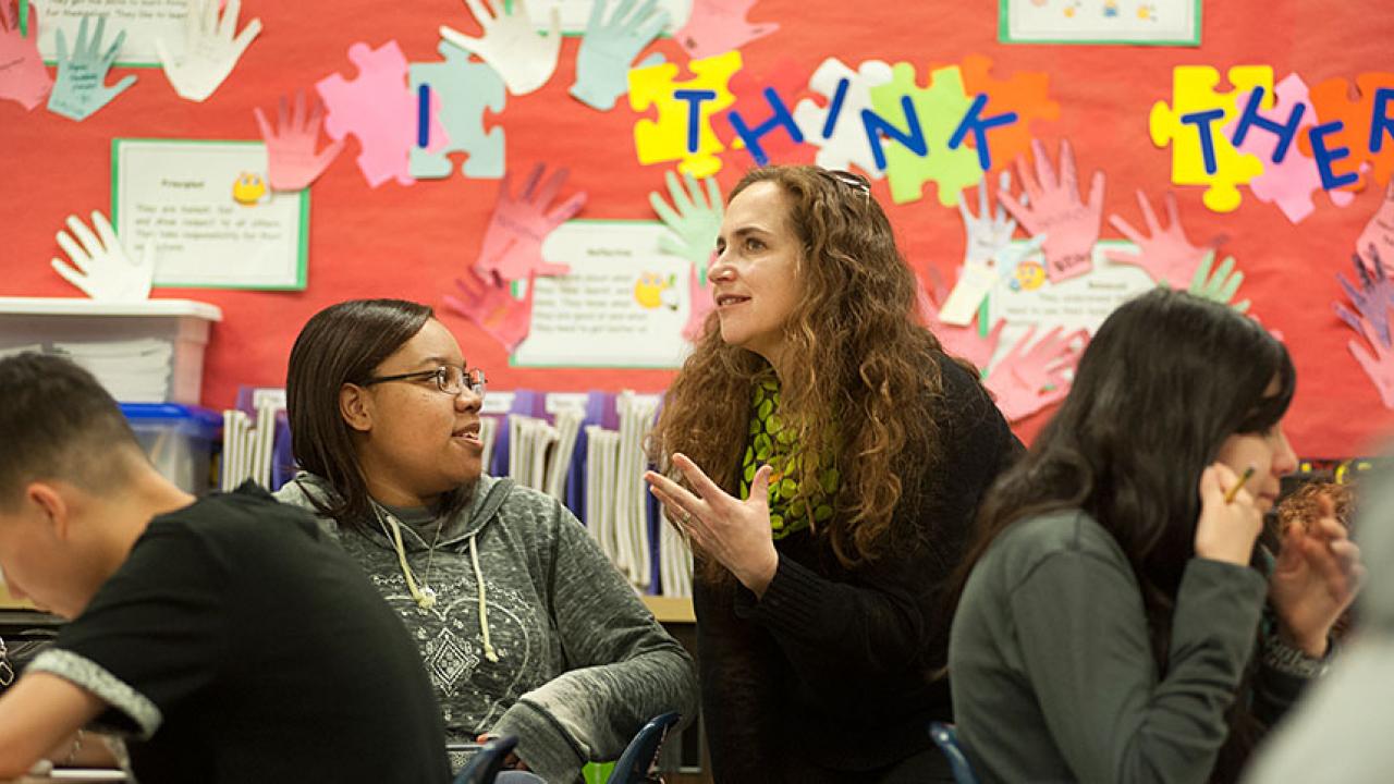 Woman in a high school classroom talking to a student.