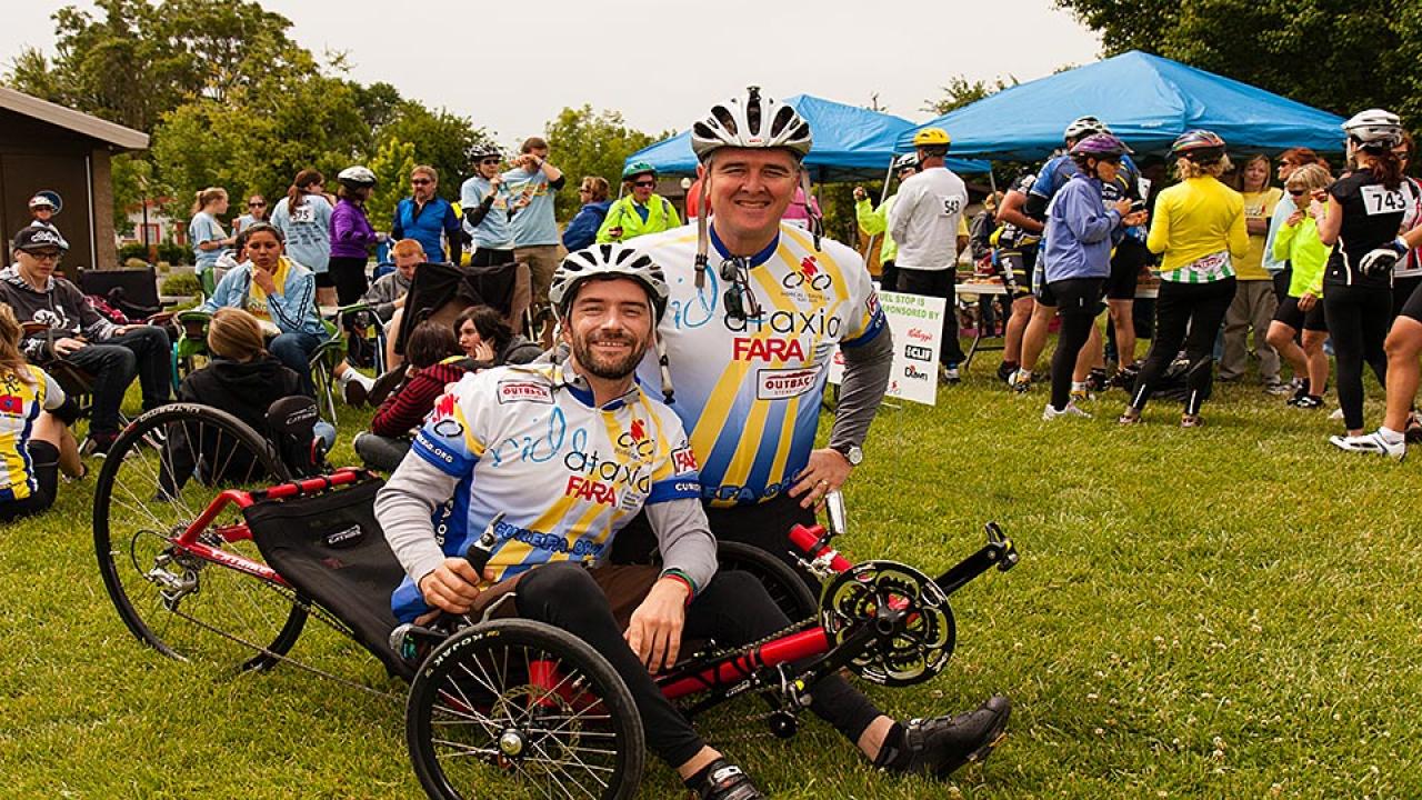 Man in bike helmet and jersey kneels next to a man seated on a recumbent bike with people and event tents behind them
