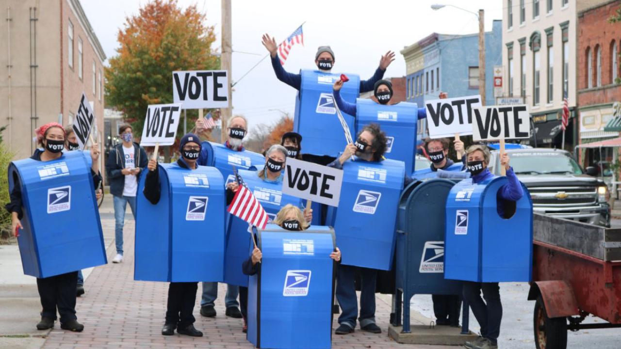 Theatre troupe in mailbox costumes, carrying "Vote" signs.