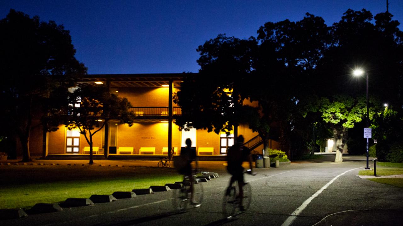 Photo: Two bicyclists on path approachining well-lit Wellman Hall at night.l