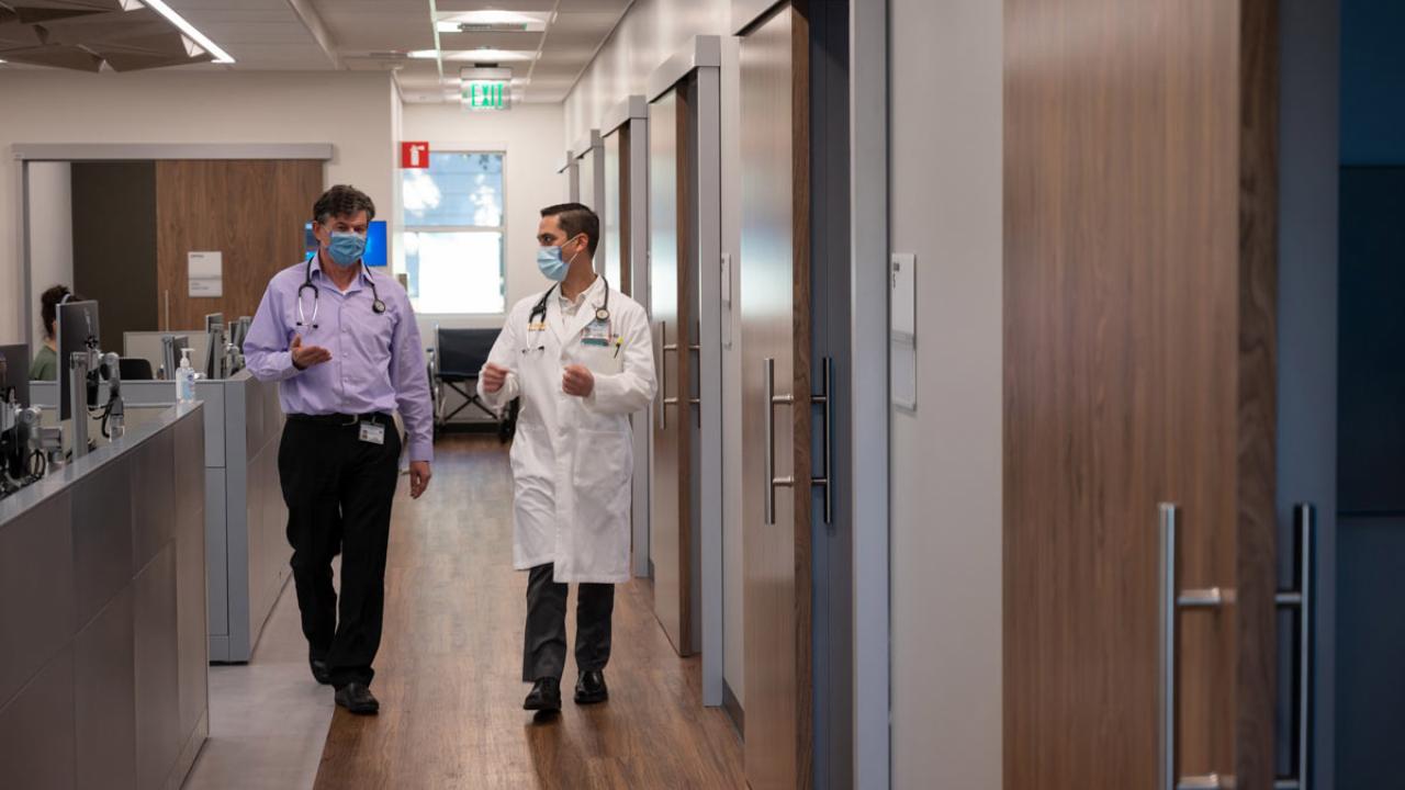 Two men (each with a stethoscope around his neck) chat as they walk in hallways, toward camera.