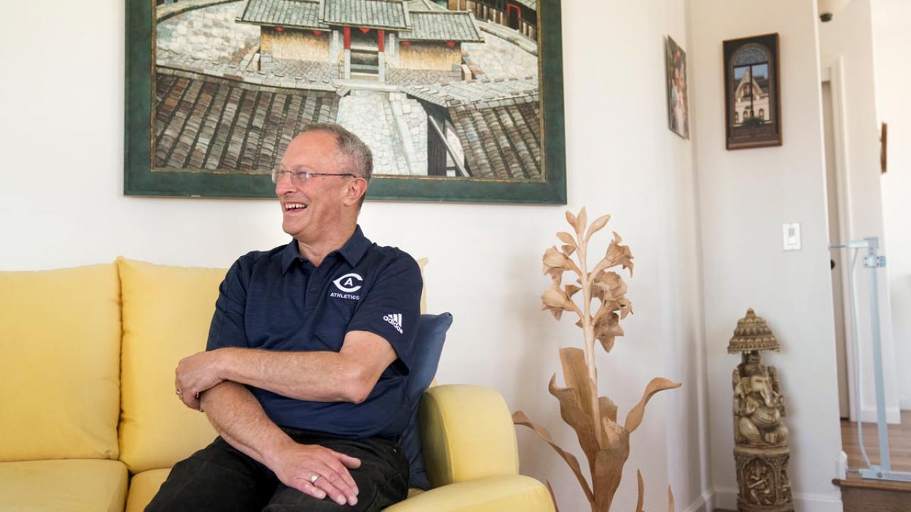 Ralph J. Hexter in Aggie polo shirt, seated on couch, under a painting.