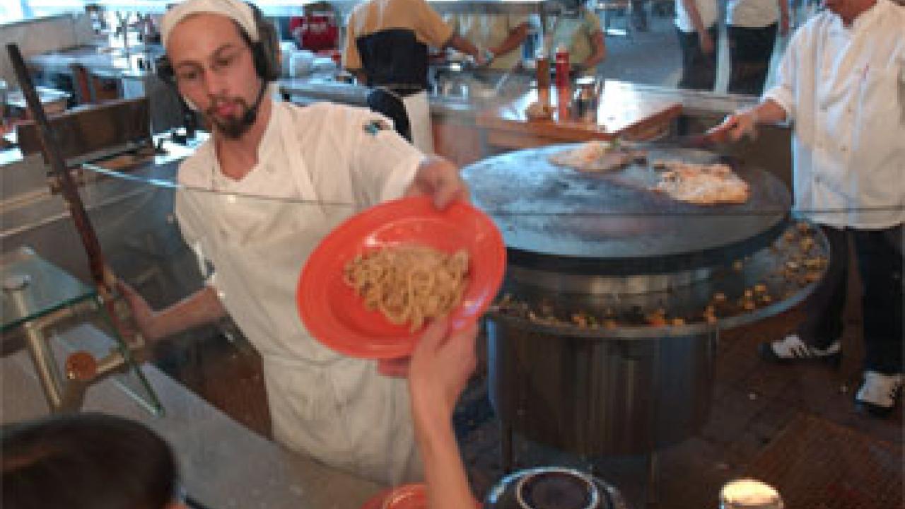 Photo: chef hands bowl of noodle to woman, with many people in the background