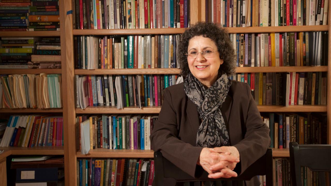 Suad Joseph, sitting at table, hands folded, in front of bookcases.