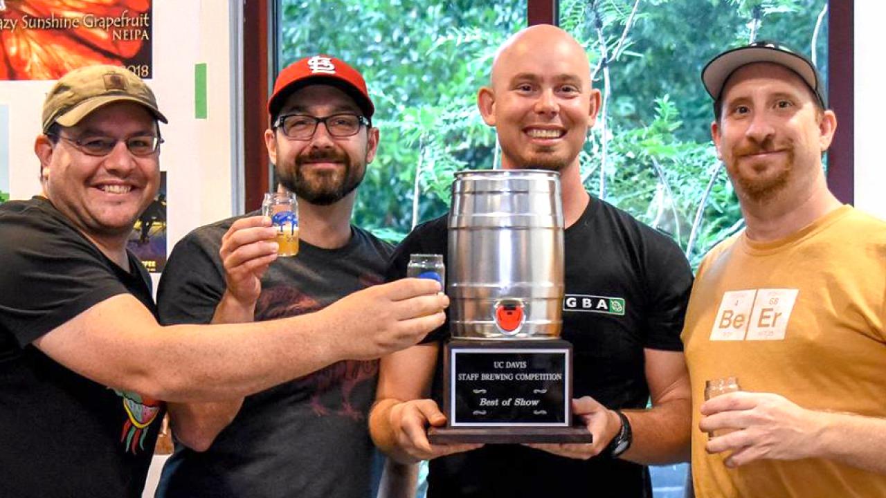 Four men holding glasses of beer and a keg trophy.