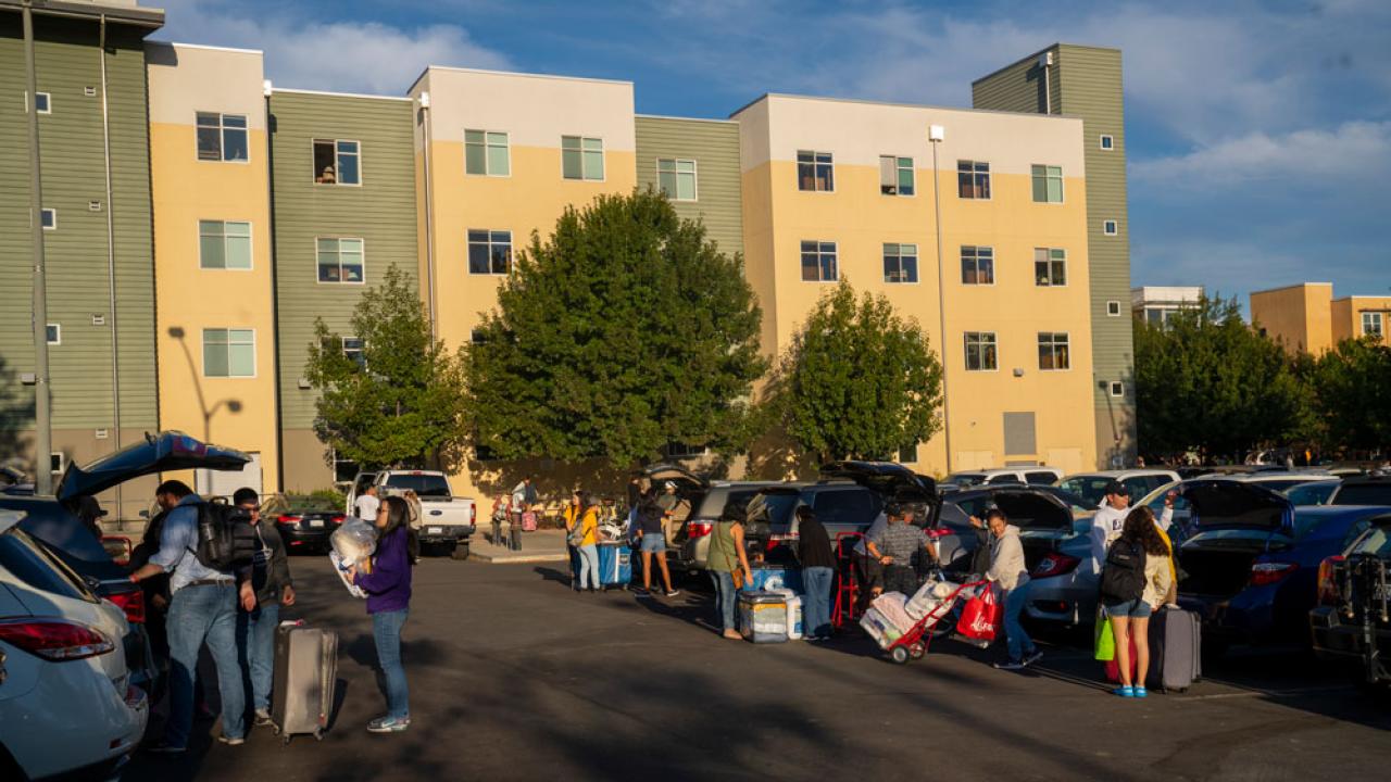 Families gather around cars, in parking lot, unloading students' belongings, with residence hall in background.